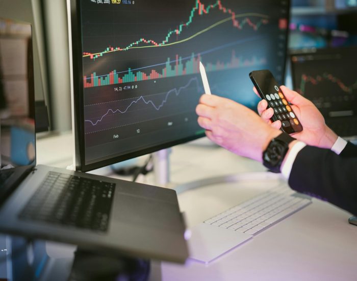 A professional analyzing financial charts on computer monitors and a smartphone, symbolizing digital finance strategies.